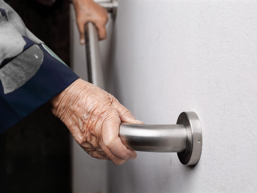Mano de adulto mayor sujetando pasamanos de acero inoxidable ergonómico montado en muro interior